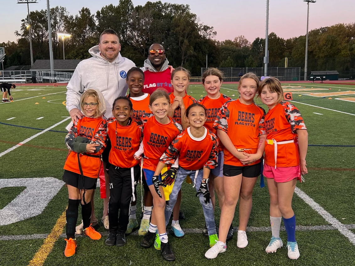 The Flag Star Bears youth flag football team in orange jerseys, smiling with their coaches on the field