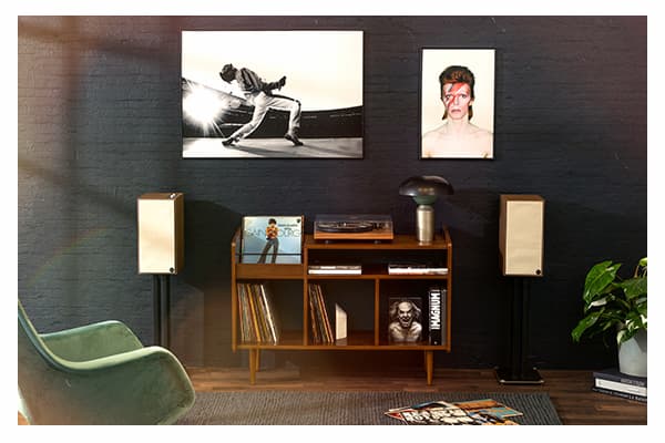 The Sevens 6.5" Powered Speakers on a white background. Pair of walnut finished bookshelf speakers with light-tan fabric grilles, posted on black-metal stands. Speakers are positioned against a black-brick wall, with pictures of celebrities , and a record cabinet and player is situated between them. 
