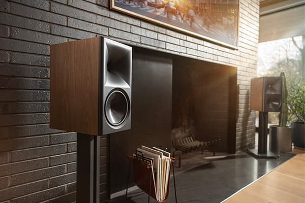 Walnut finished Klipsch Fives II speakers on dark metal stands sit next to a fireplace in a bright living room