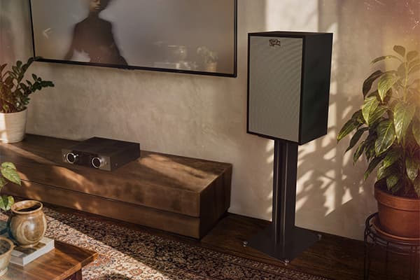 Black Klipsch Rebellion speaker, on a speaker stand, in a dimly lit living room.