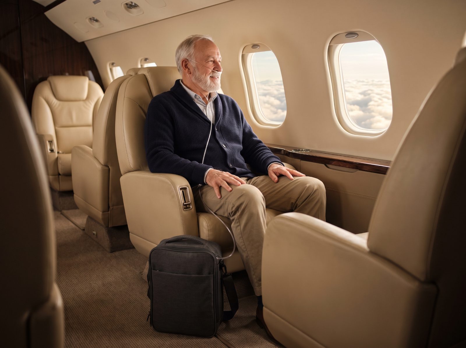 An older man sits at an airplane window with a portable oxygen concentrator in a carry bag at his feet.