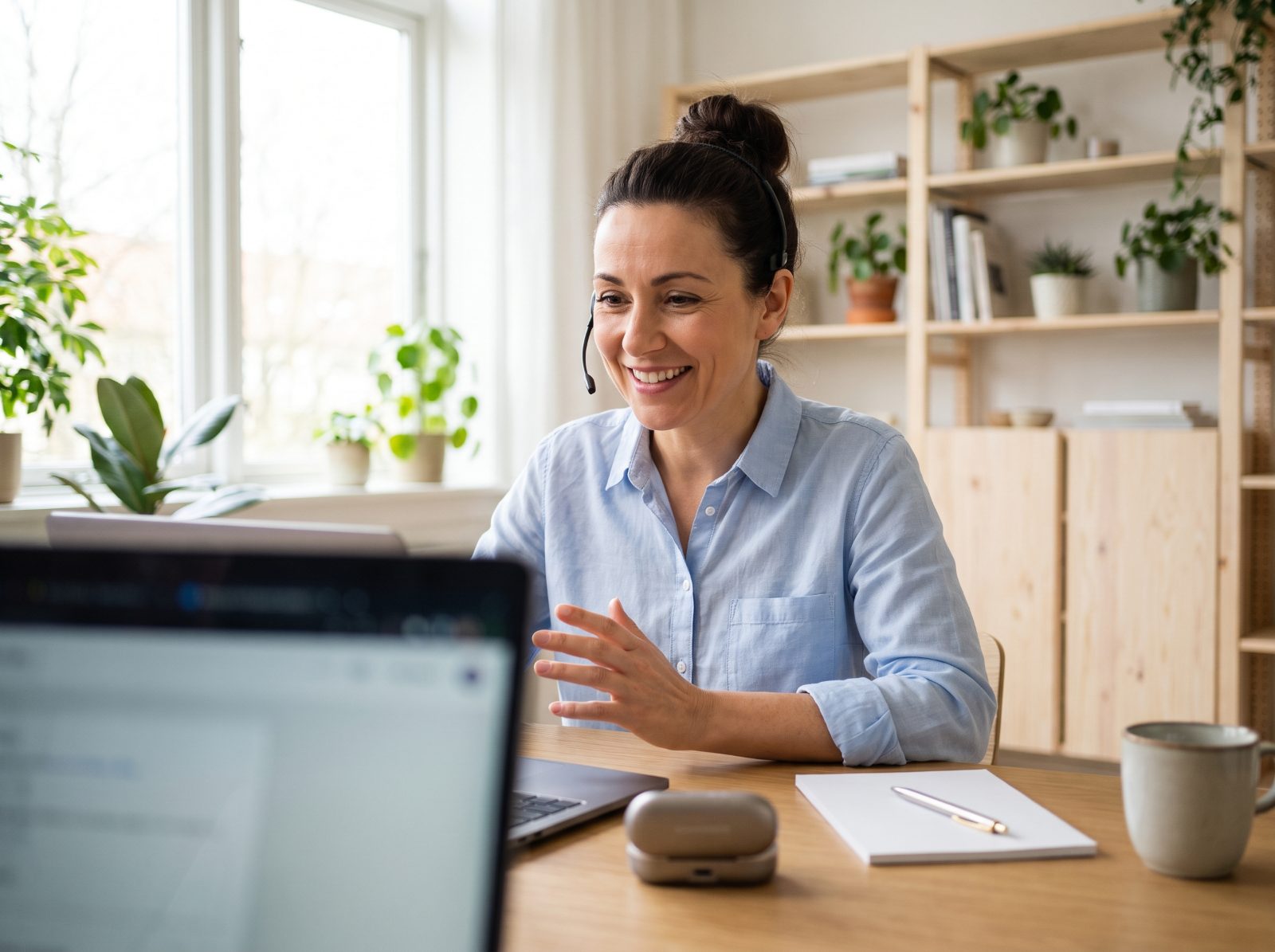 A smiling audiologist with a headset talks with a client on a video call in a bright office.
