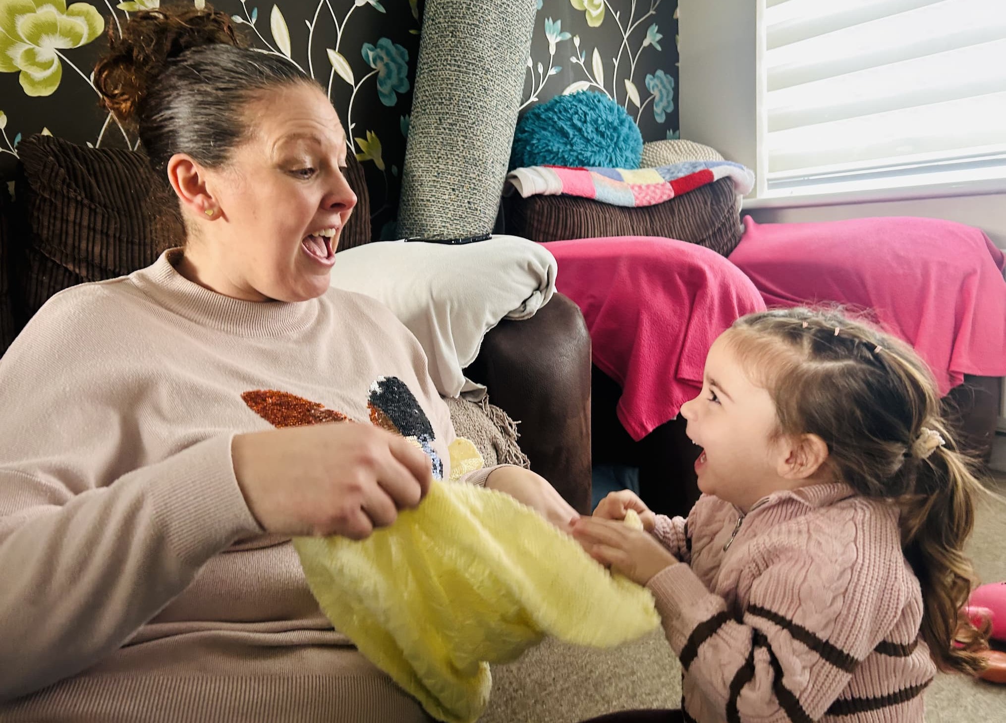 Childminder Kerry Keenan playing in her home with a young girl, both laughing as they play with a yellow fluffy blanket