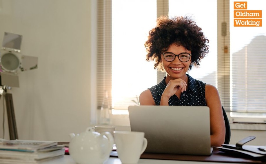 A person wearing spectacles smiling while seating at a laptop computer with a cup and teapot in front of them