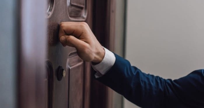A man wearing a dark suit and white shirt knocking on a dark wooden door