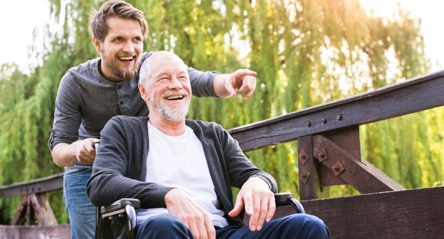 A man smiling and pointing as he stands behind an older man who is seated in a wheelchair