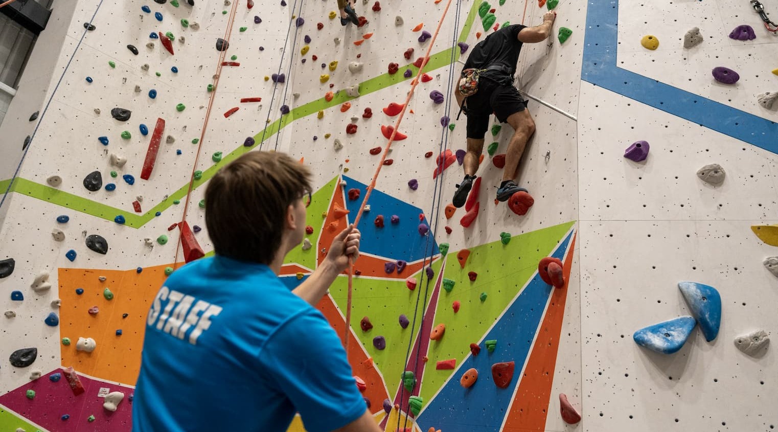 A man scaling a climbing wall at Summit Up as a member of staff holds the ropes and looks on