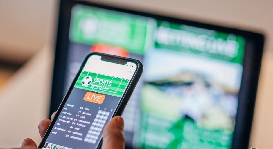 A hand holding a TV remote displaying the words 'Sports Betting Live'