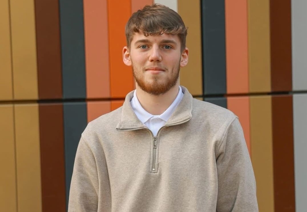 A smiling Jadon wearing a beige zip-next detail sweatshirt over a white polo shirt standing in front of a striped beige, navy, burnt orange and terracotta backdrop