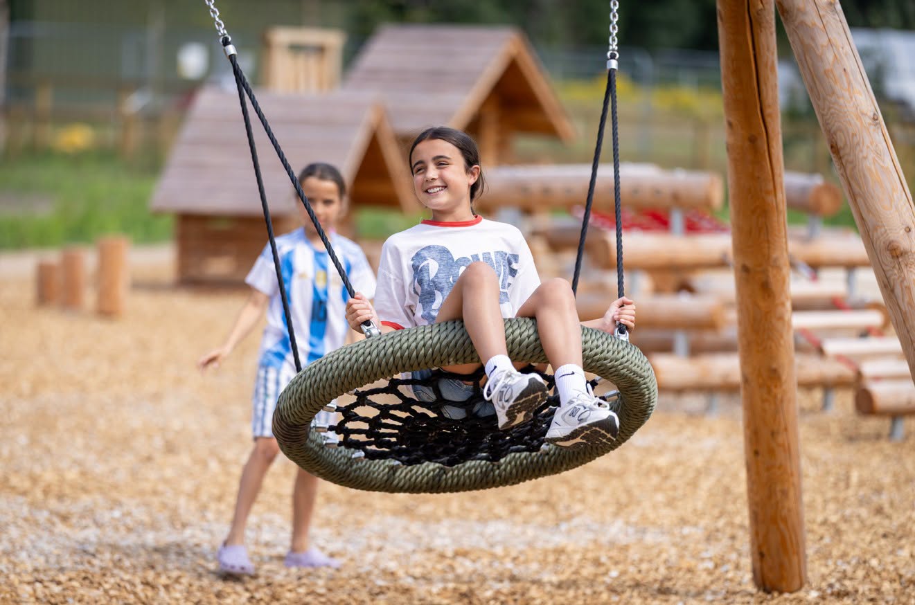 Two children playing in a playground. One is smiling as they sit on a swing pushed by the other child 