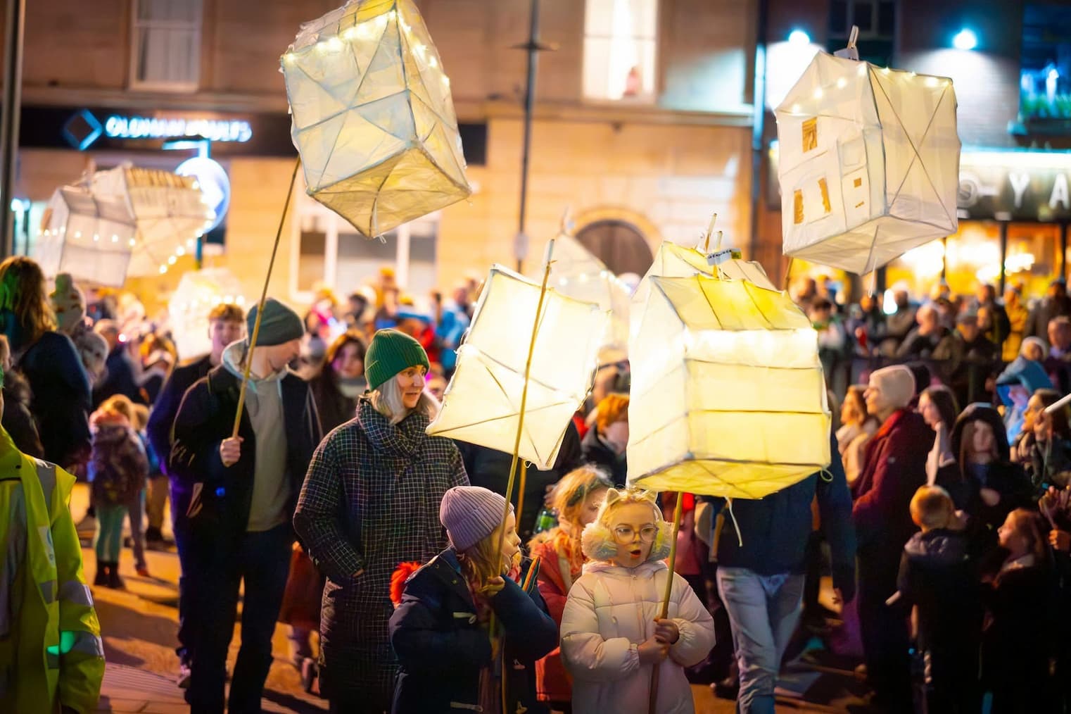 Adults and children wearing warm clothing and hats as they carry glowing lanterns through Oldham town centre 