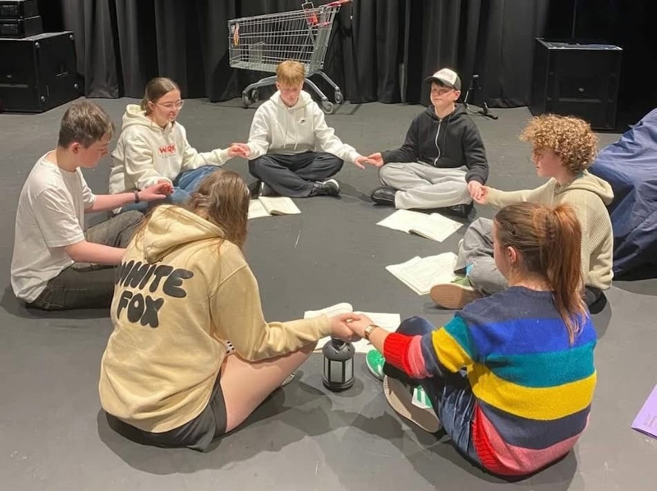 Seven young people seated in a circle on the floor holding hands