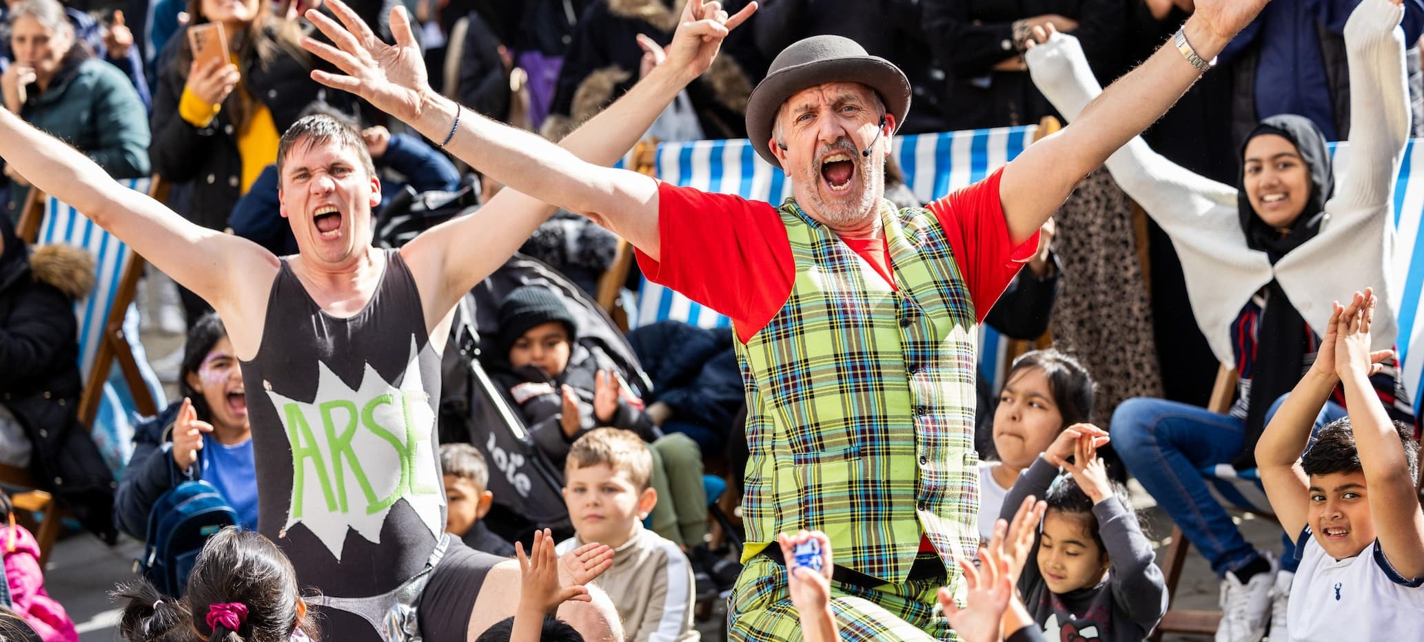 Street performers wearing eye-catching outfits - colourful vests and a bowler hat - as they raise their arms in the air mid-performance as a crowd of children follow