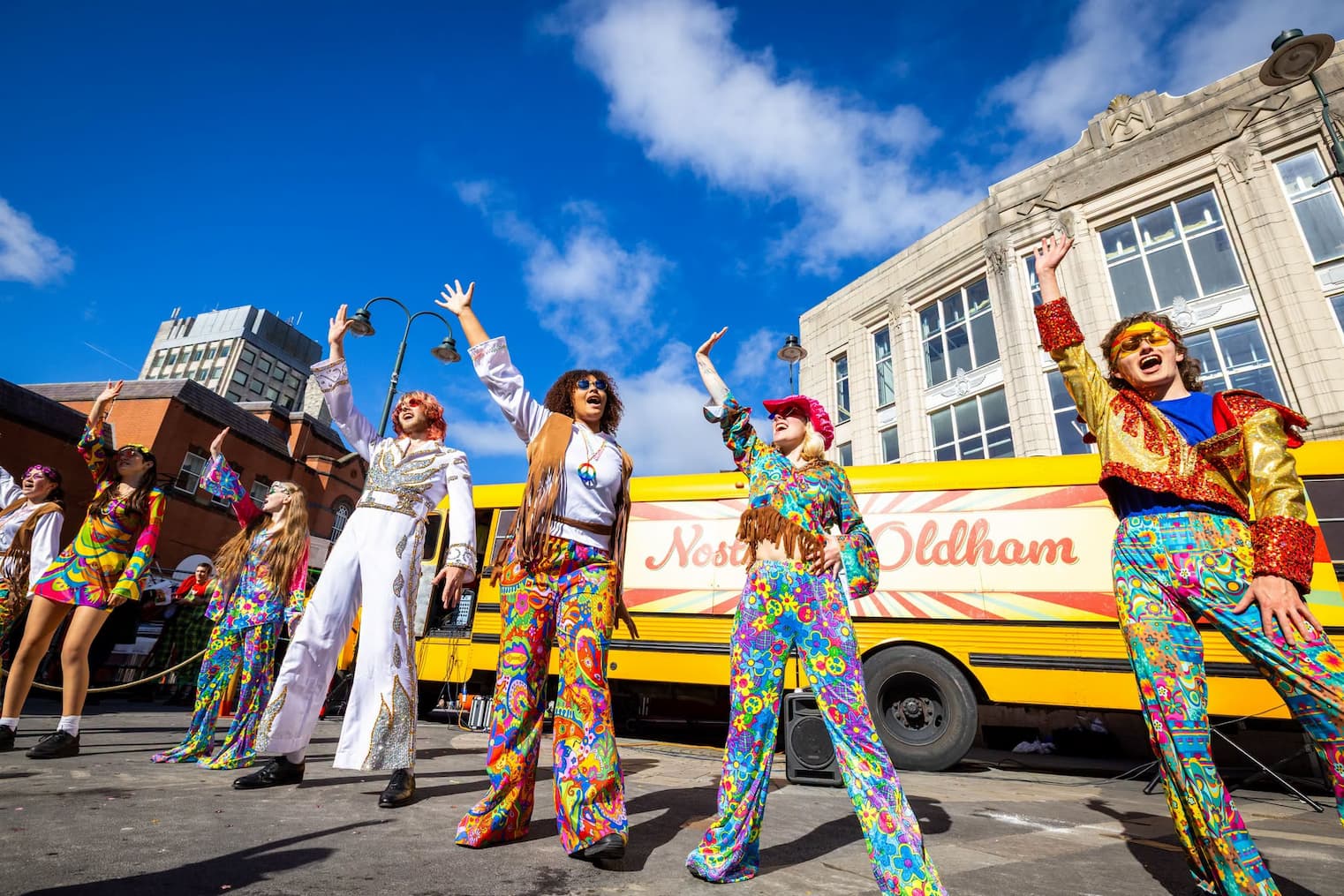 A group of 70s retro performers in floral outfits and flares in front of a yellow Nostalgic Oldham bus