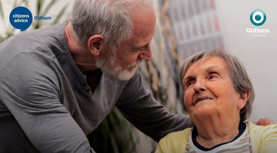 An elderly man and woman smiling at each other, the man's arm is around the woman's shoulders