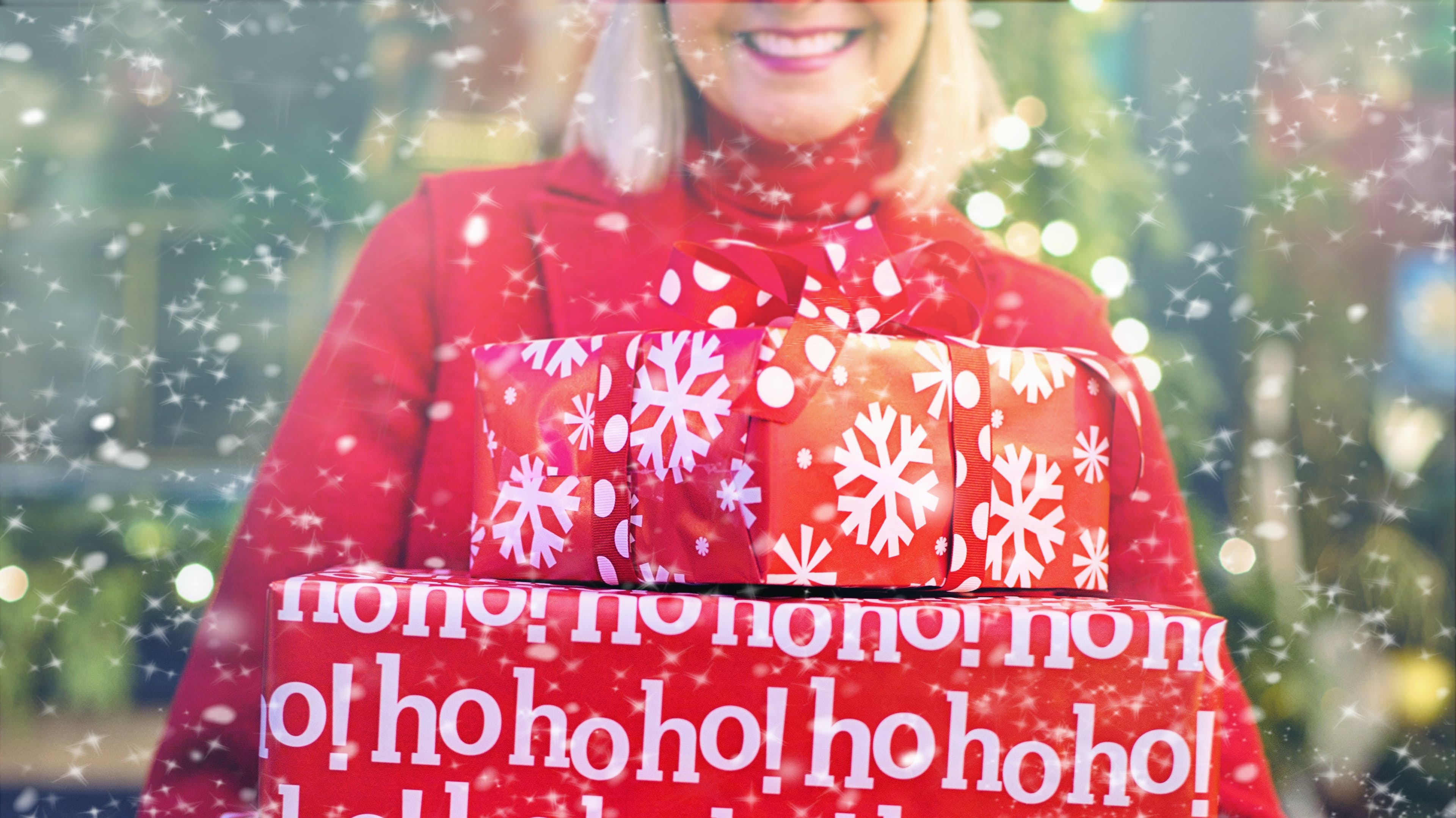 Image of woman carrying Christmas gifts wrapped in red and white patterned paper as snow falls
