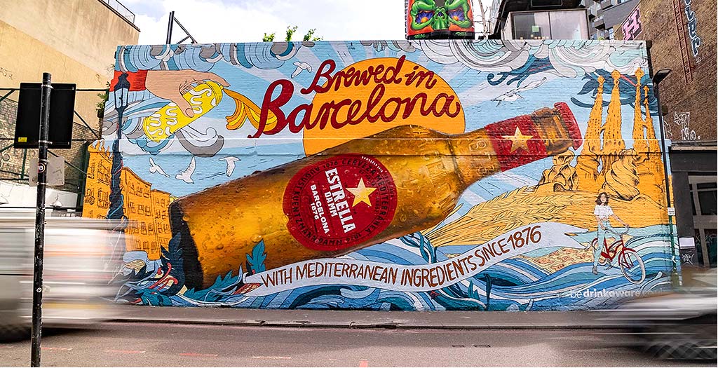 The same drinks artwork hand-painted at building scale across a brick wall beside a road
