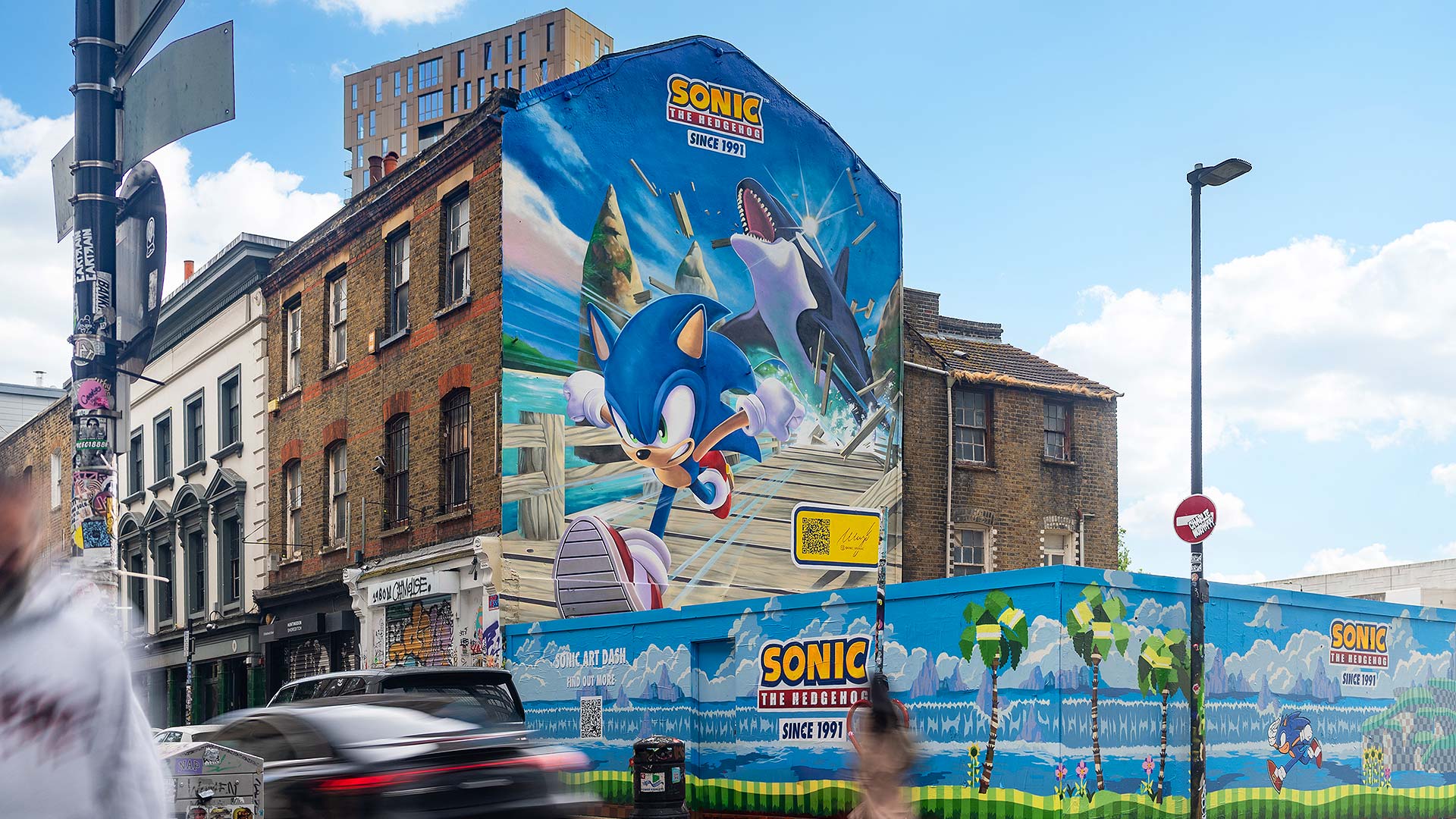 A hand-painted video game character covering the end wall of a building on a street corner, with a painted hoarding running along the pavement below
