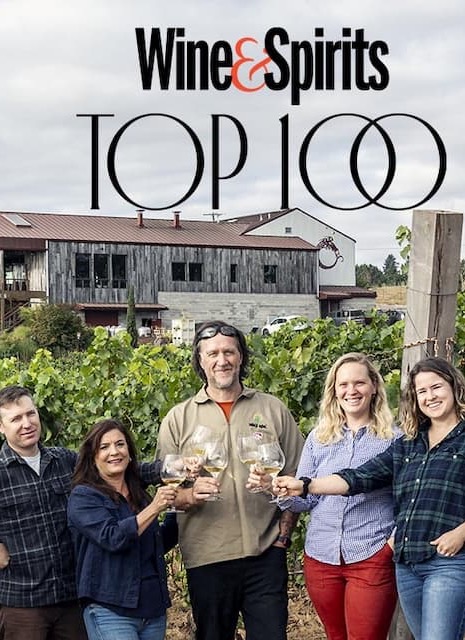 Magazine-style image labeled “Wine & Spirits Top 100,” showing a group of people standing in a vineyard in front of a rustic winery building, raising glasses of white wine together.