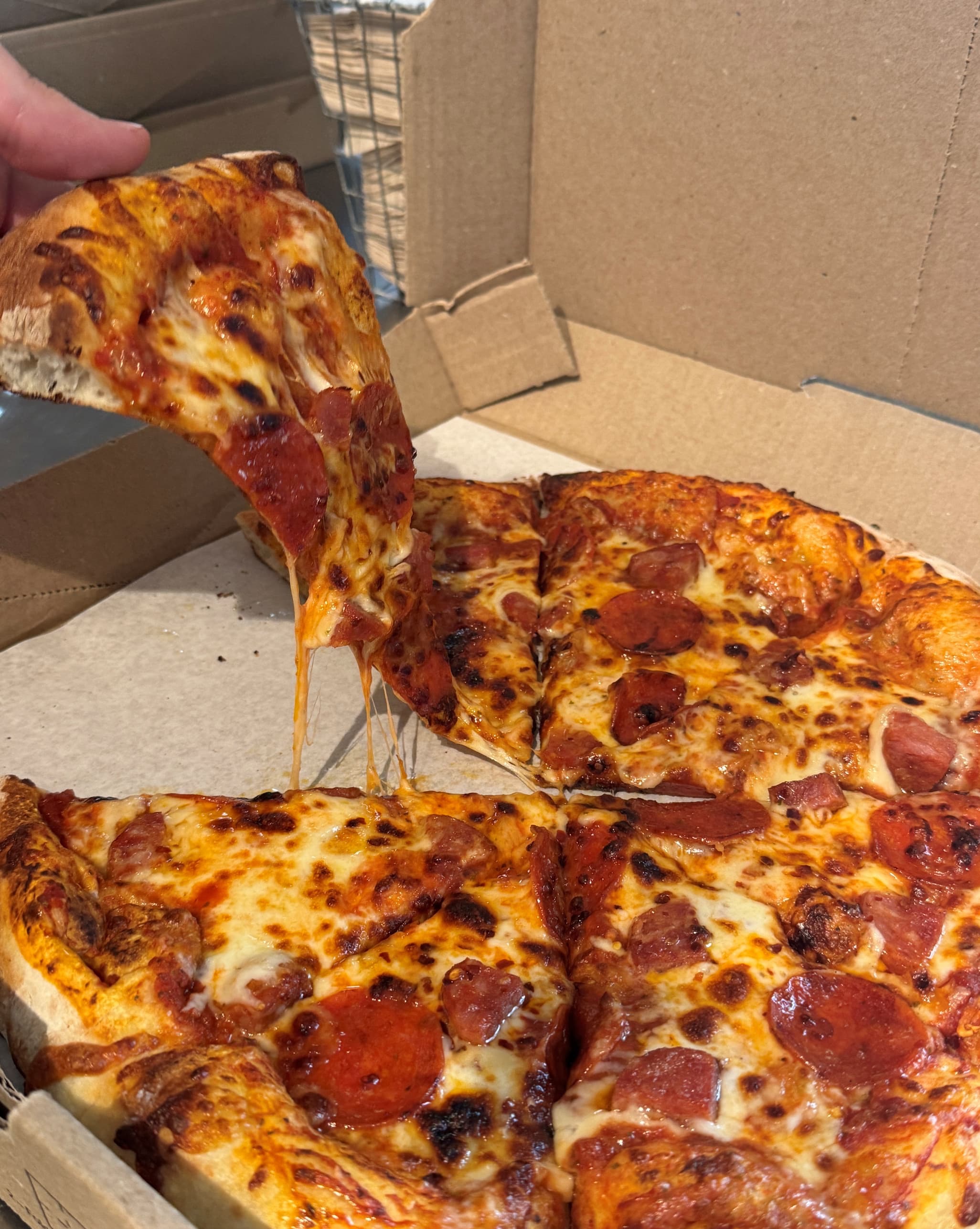 Hand lifting a slice of pepperoni pizza from a cardboard box, with melted cheese stretching between slices, blistered crust, and browned, bubbling cheese topped with pepperoni.