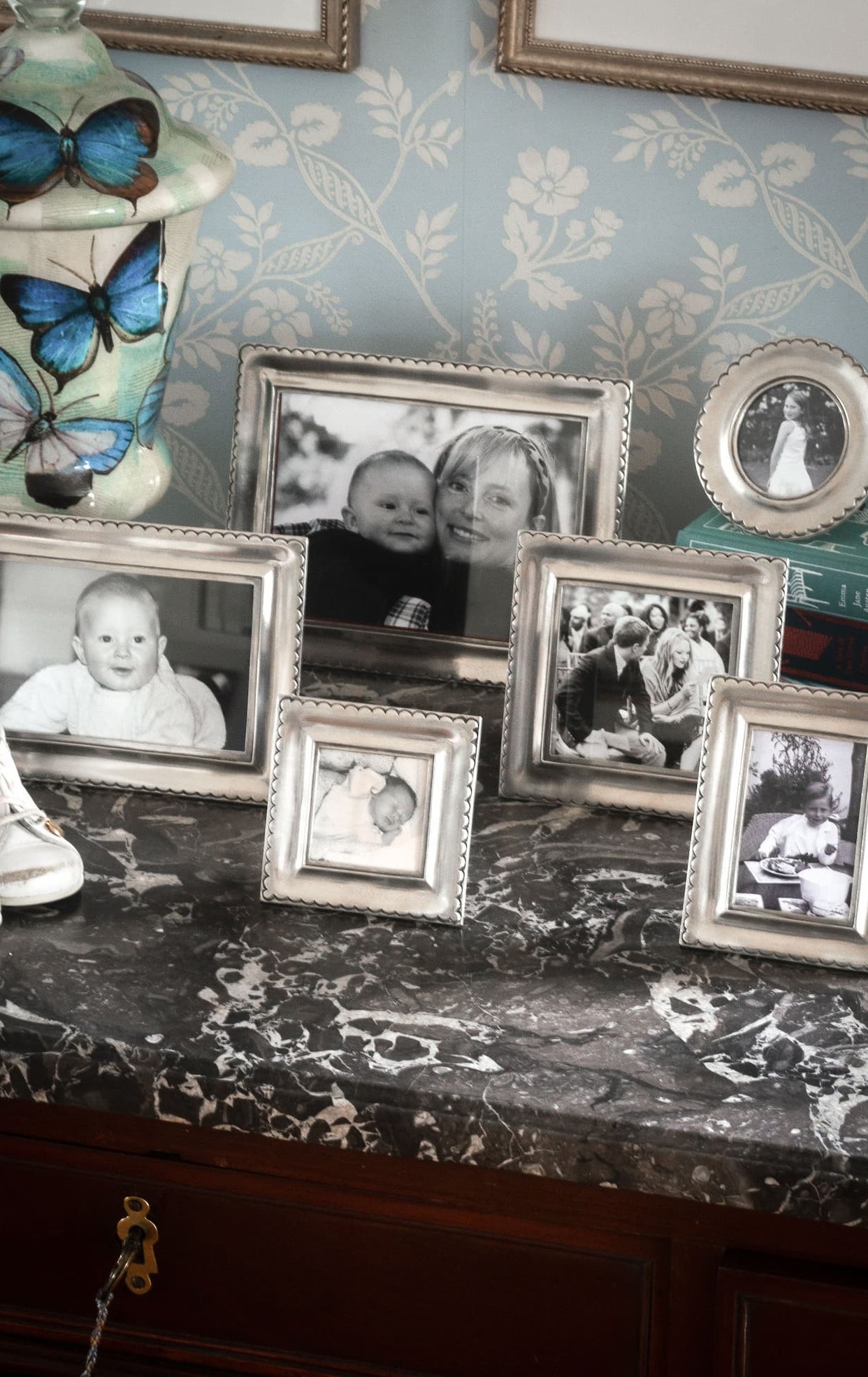 Collection of silver picture frames in assorted sizes displaying black-and-white family photographs on a marble-topped console, accented by a butterfly-patterned vase. Fearrington Village.