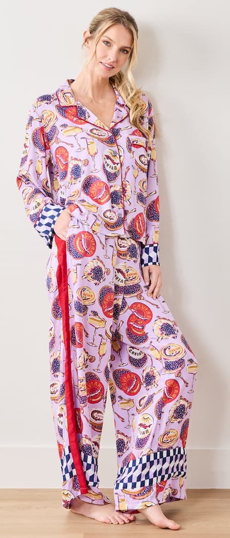 Woman wearing a lilac pajama set with playful food and drink illustrations, contrast patterned cuffs, and wide-leg pants, standing barefoot against a light wall with one hand in her pocket.