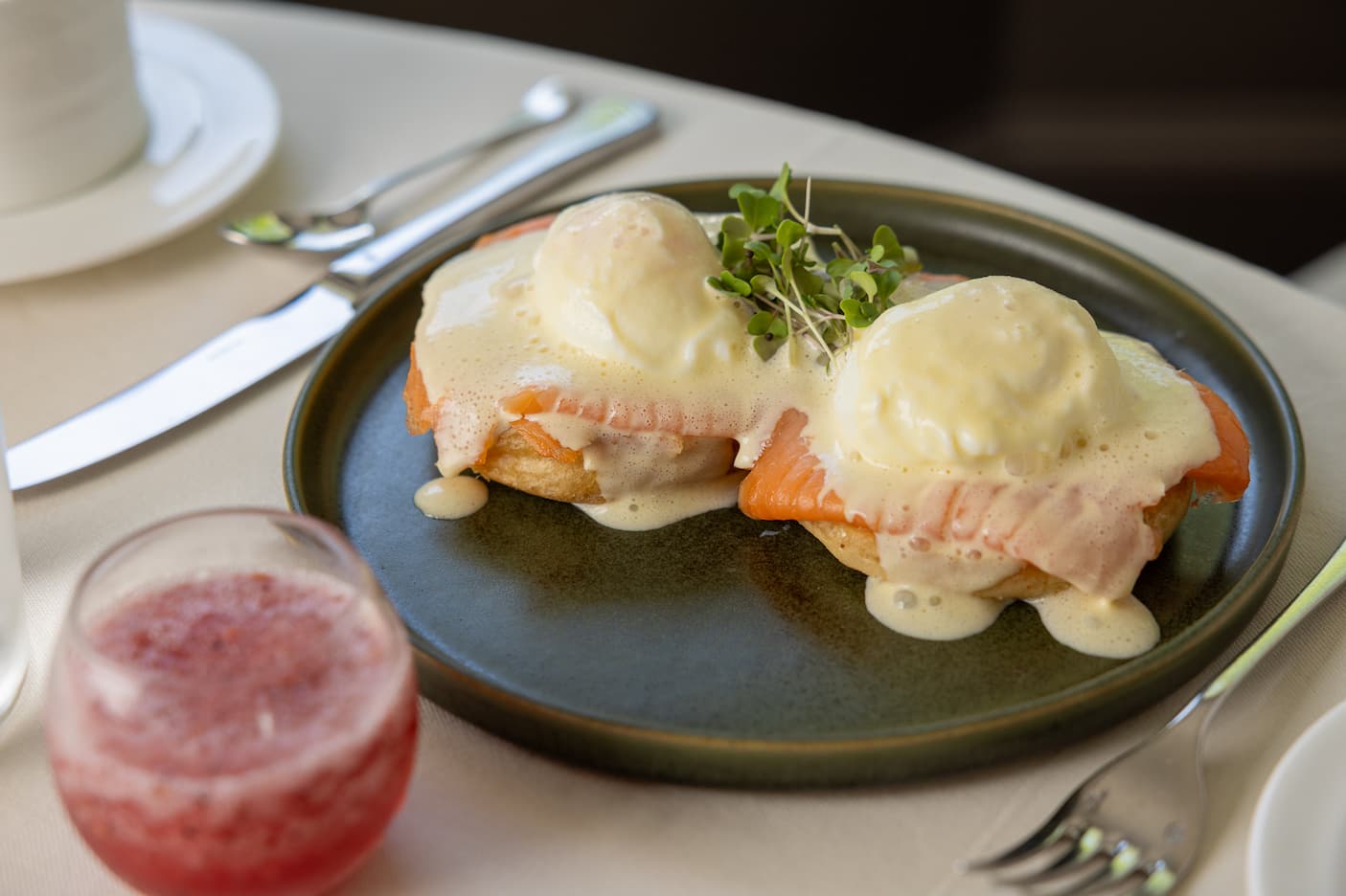 Smoked salmon eggs Benedict topped with silky hollandaise and microgreens, served on a dark ceramic plate beside a sparkling pink brunch cocktail in an elegant dining setting.
