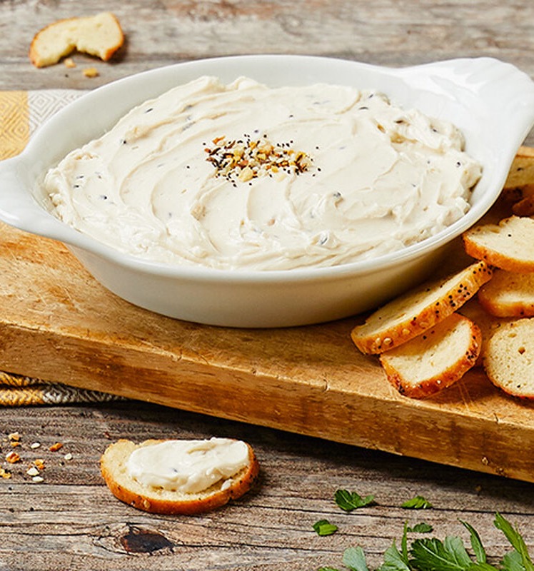 Creamy white dip in a shallow serving dish, topped with seasoning, set on a wooden board with sliced baguette rounds for spreading.