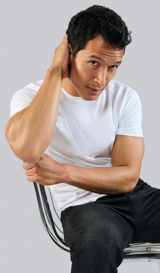 A man in a fitted white T-shirt sits on a chair against a light background, one hand reaching behind his head while he looks slightly upward.
