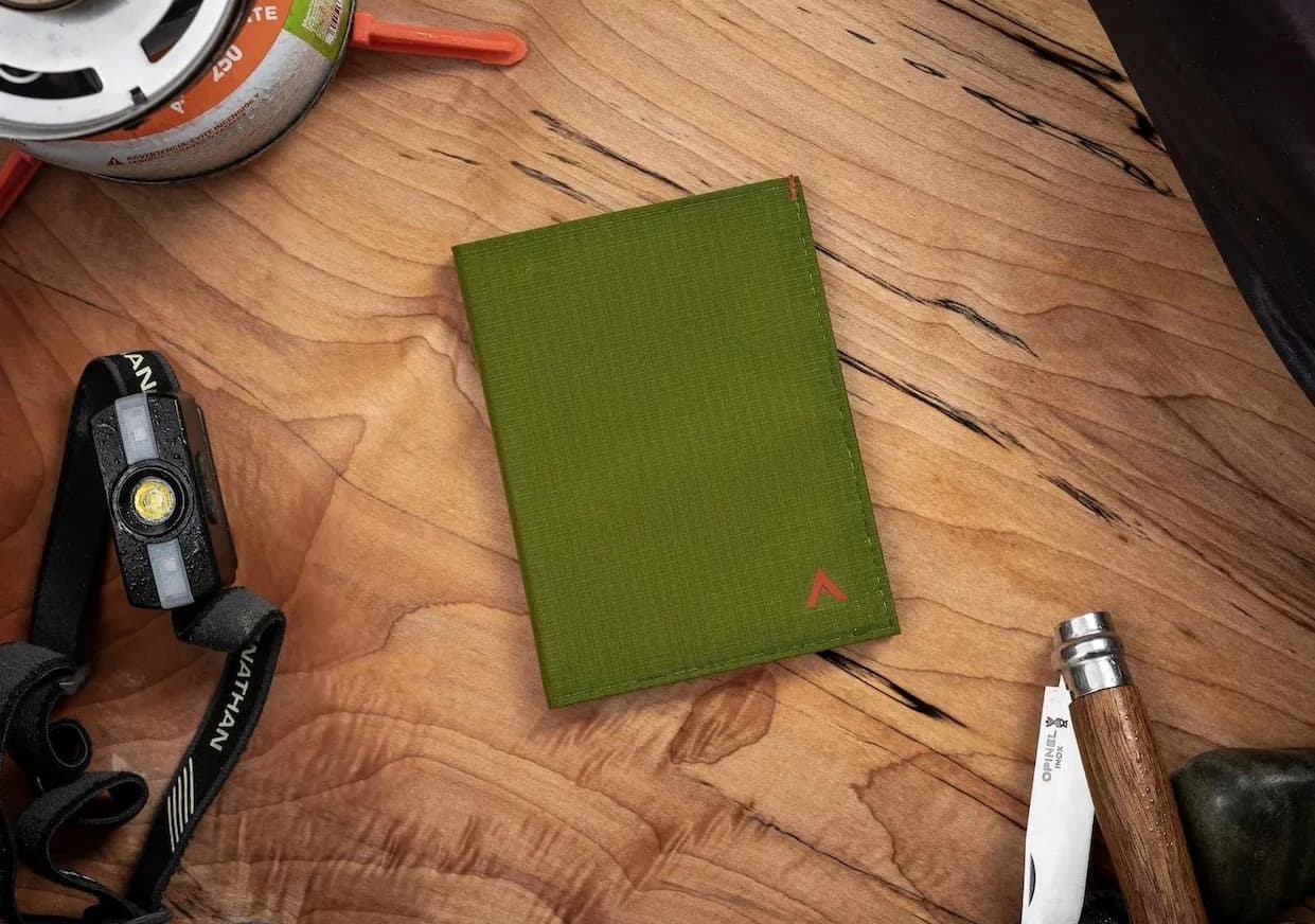 An olive green travel wallet rests on a wooden tabletop surrounded by outdoor gear, including a headlamp, camp stove fuel canister, and folding knife, highlighting its rugged, adventure-ready design.