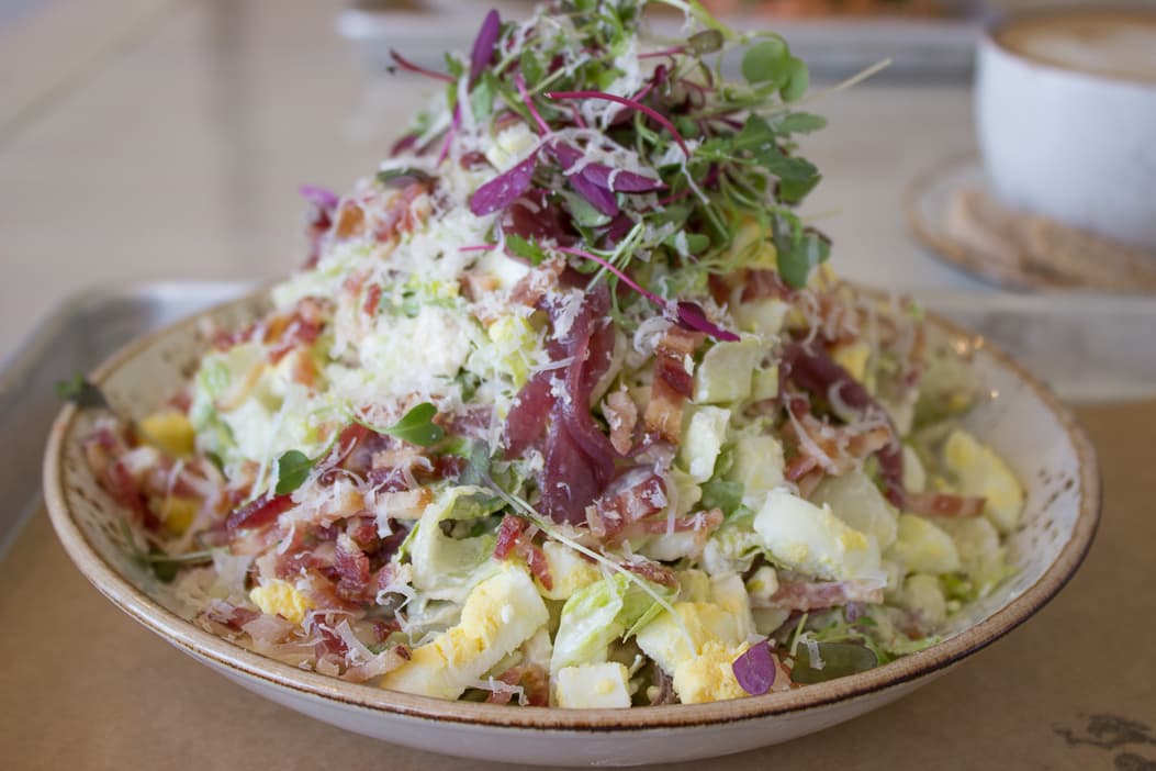 Heaped chopped salad topped with bacon, hard-boiled egg, shaved cheese, and microgreens in a shallow bowl, with a cup of coffee blurred in the background.