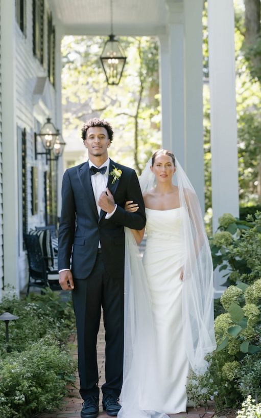 Bride and groom walking arm in arm along a garden-lined path, the bride in a simple white gown with a long veil and the groom in a black tuxedo with a boutonniere, outside a columned house with lanterns.