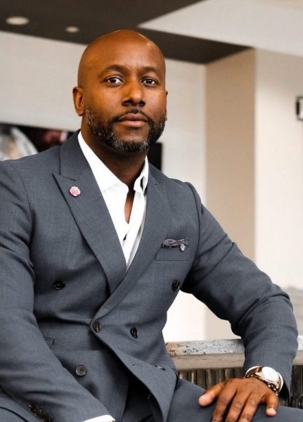 A man in a gray suit sits confidently on a chair, looking at the camera with a calm expression in a modern indoor setting. Fearrington Village