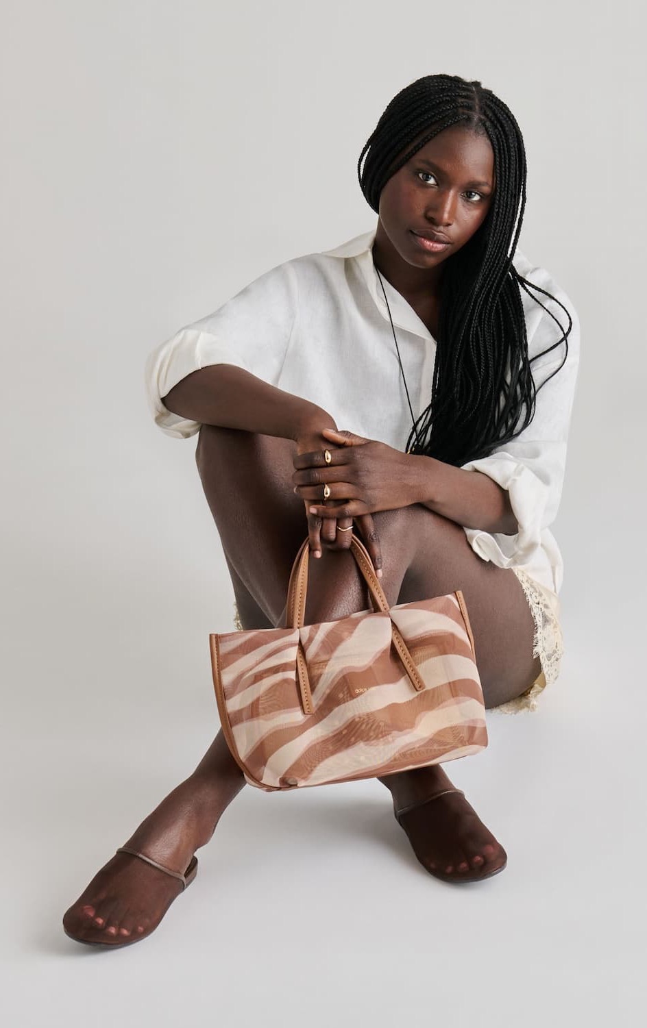 Studio portrait of a model seated on the floor against a light gray background, wearing an oversized ivory button-down shirt, cream lace shorts, and translucent brown mesh slide flats. She holds a small structured tote featuring a soft, abstract zebra-inspired pattern in warm beige and blush tones with tan handles and trim. The neutral palette and relaxed styling create a modern, understated look.