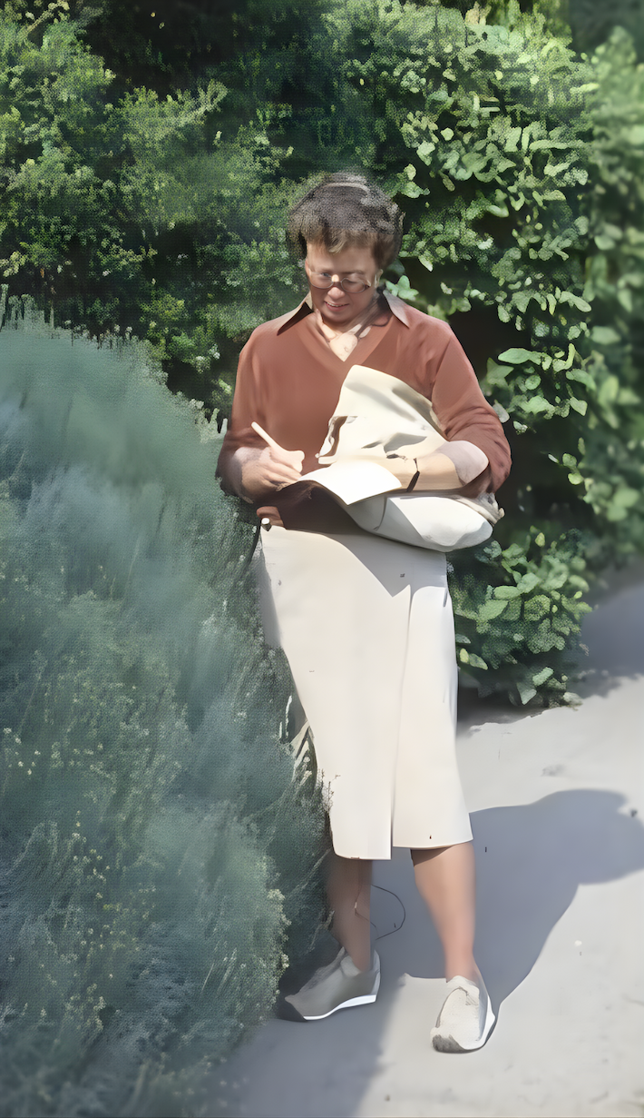 Jenny Fitch standing on a garden path in bright sunlight, writing in a notebook while leaning against soft green shrubs, wearing a rust-colored blouse, light skirt, and sneakers.