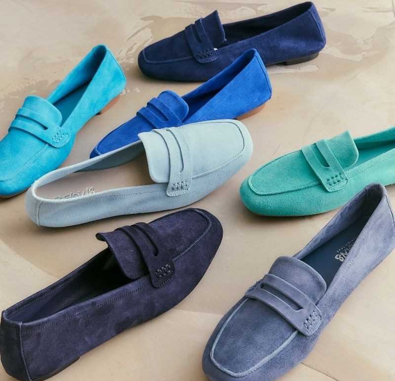 Seven suede loafers in various shades of blue, teal, and green are arranged on a light beige surface, with some shoes facing forward and others turned to the side. Fearrington Village