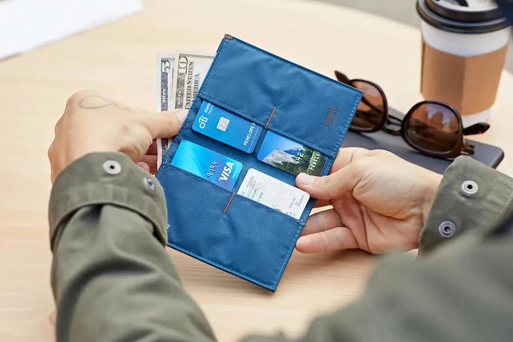 A person holds an open blue travel wallet featuring multiple card slots and a cash pocket with U.S. currency. Sunglasses, a takeaway coffee cup, and a tablet rest on the table nearby, suggesting travel or everyday organization.