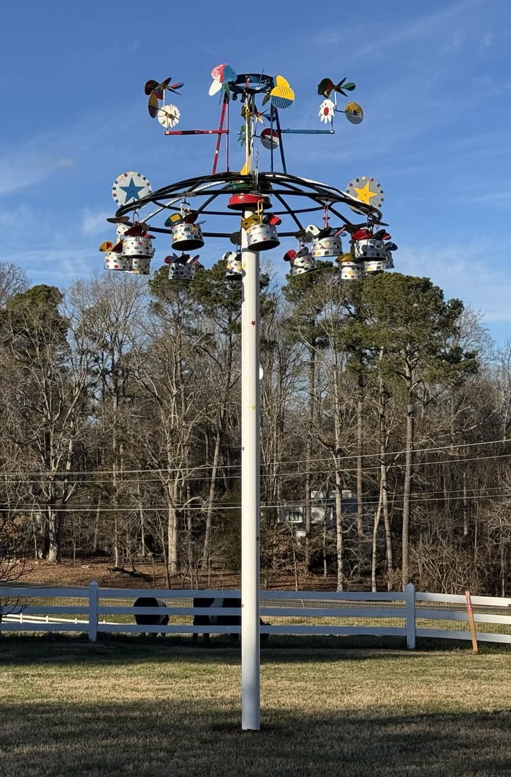 Tall whirligig wind sculpture on a pole in a grassy field with trees and a white fence behind it.