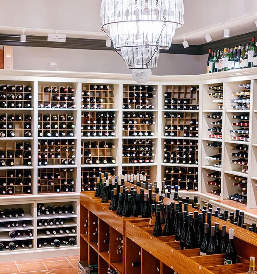 A beautifully organized wine shop features floor-to-ceiling shelves filled with bottles from around the world. Wooden display tables in the center showcase additional selections, while a large crystal chandelier hangs overhead, casting a warm glow across the space. The room feels elegant, inviting, and designed for leisurely browsing and discovery.