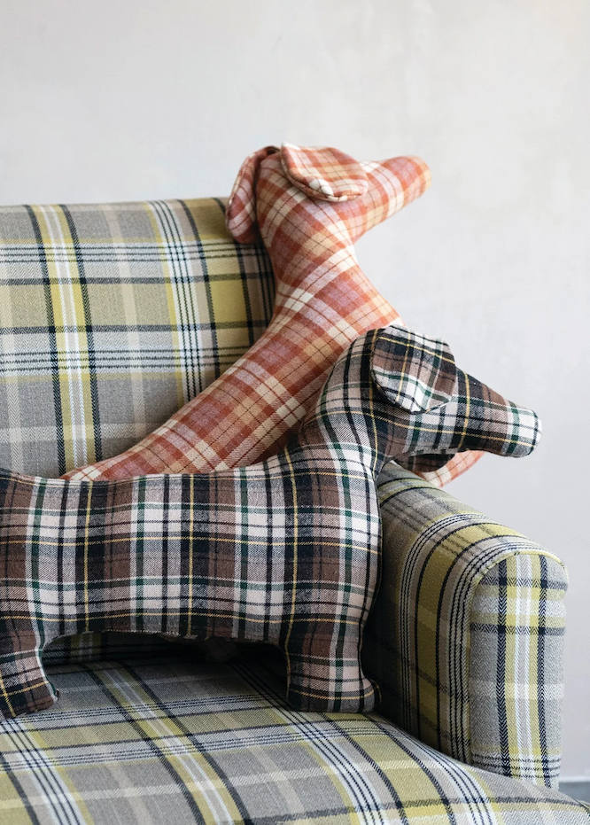 Two plaid fabric dog-shaped decorative pillows rest on a matching tartan armchair, featuring soft muted tones of rust, brown, cream, and sage green. The cozy scene blends playful charm with classic cottage-inspired style.