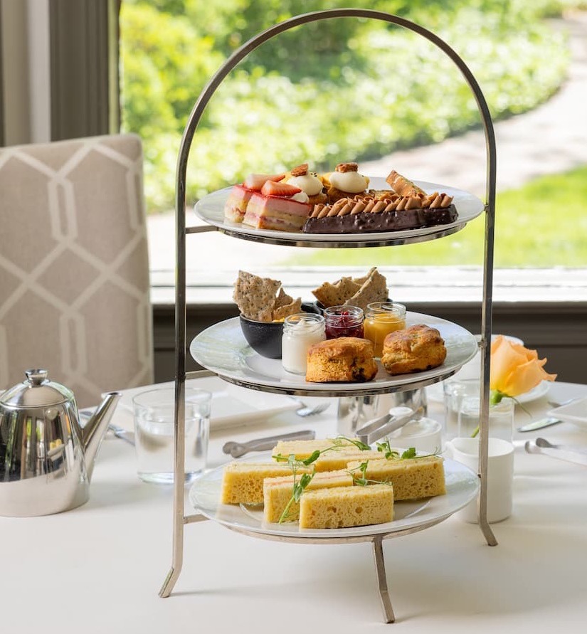 Elegant three-tier afternoon tea stand featuring finger sandwiches, fresh scones with preserves and cream, and an assortment of pastries and desserts, set on a white tablecloth beside a silver teapot and garden-view windows.