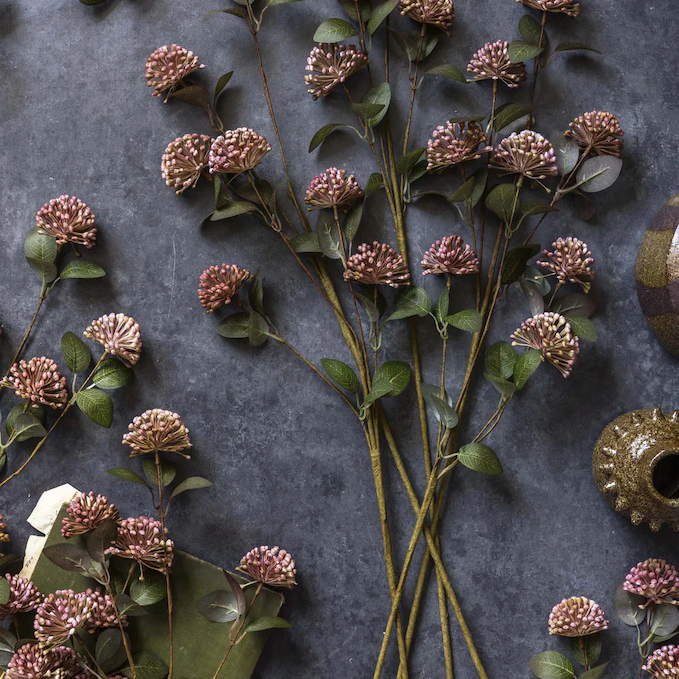 Muted mauve faux floral stems arranged across a moody charcoal surface with scattered greenery and decorative objects. The textured blooms and earthy tones create an elegant, artful botanical flat lay.