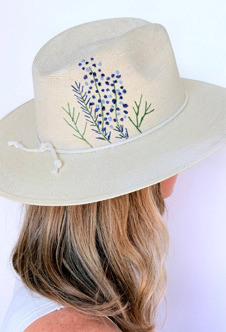 Back view of a handwoven ivory straw hat featuring delicate blue and green floral embroidery on the crown, shown on a woman with shoulder-length blonde hair against a soft white background. Fearrington Village.