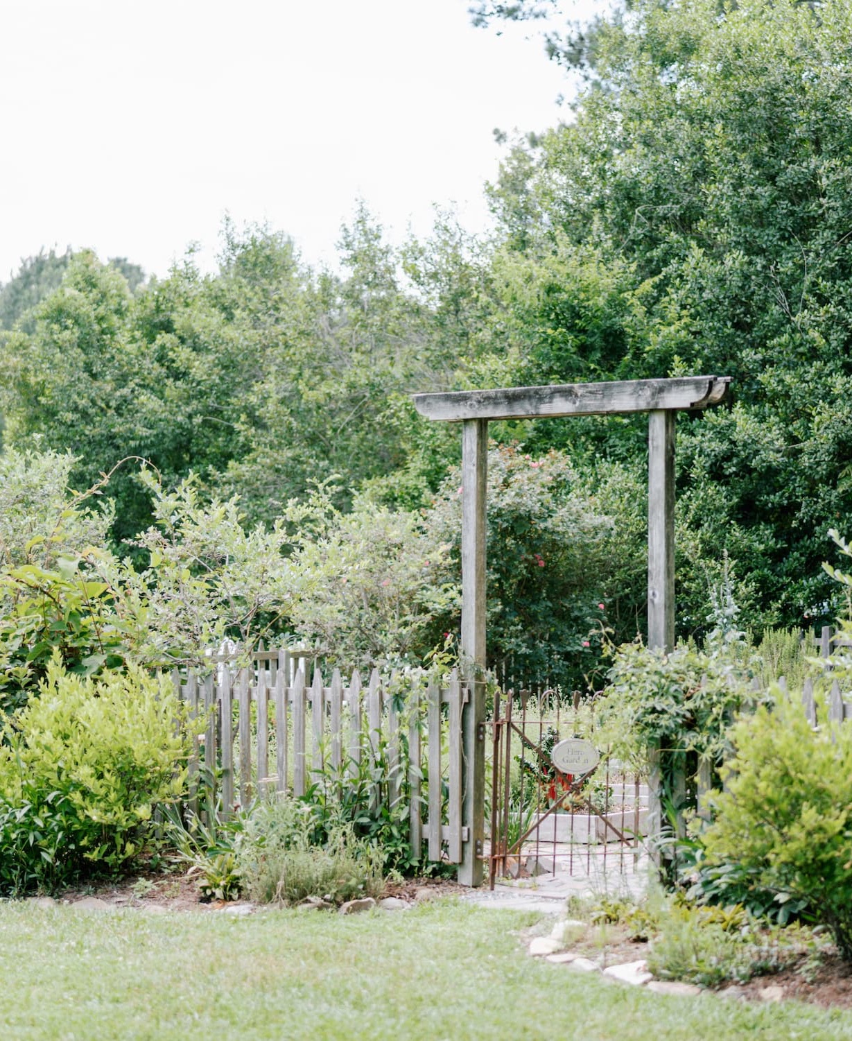 Lush garden entrance framed by a weathered wooden arbor and a low picket fence, with a small metal gate leading inside. Surrounded by layered greenery, flowering shrubs, and soft lawn, creating a quiet, tucked-away garden setting.