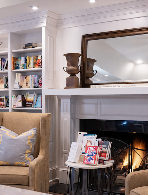 Cozy literature room at McIntyre's Books, with built-in white bookshelves filled with books, a lit fireplace, upholstered armchairs, and a mirror above the mantel.