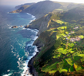 A dramatic coastal landscape shows steep green cliffs dropping into the ocean, with waves breaking along the rocky shoreline and patchwork fields and small homes spread across the sunlit hills above.