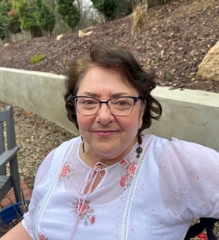 A woman wearing glasses and a light embroidered blouse sits outdoors, smiling softly against a natural, garden-like backdrop.