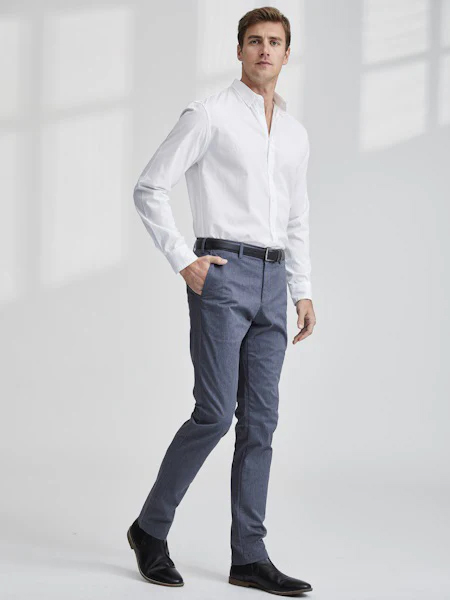 Full-length studio portrait of a man wearing a crisp white button-down shirt with tailored blue trousers, a black belt, and black leather shoes. He stands against a soft gray background with one hand in his pocket, styled in a clean, modern, and polished look.