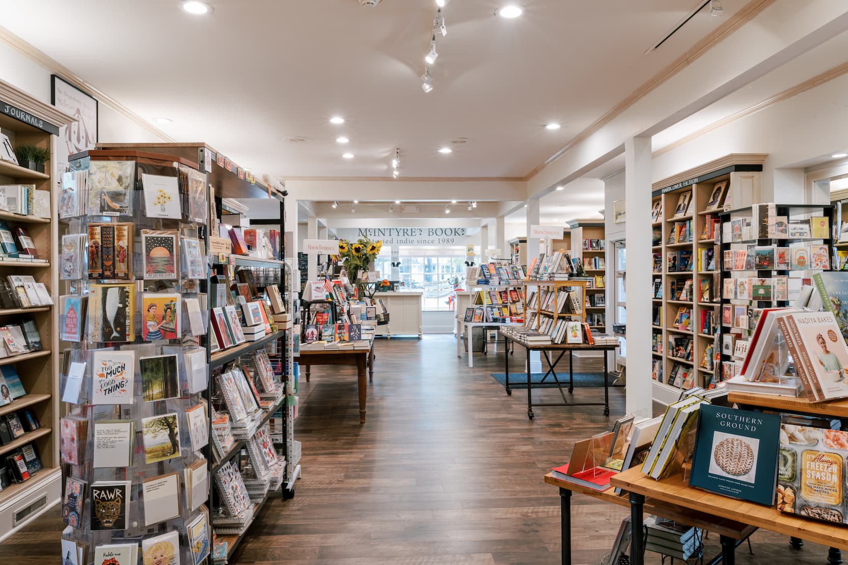Bright, inviting bookstore interior with wooden floors, neatly arranged shelves of books, greeting cards, and gifts, plus central display tables; signage for “McIntyre’s Books” visible toward the back.