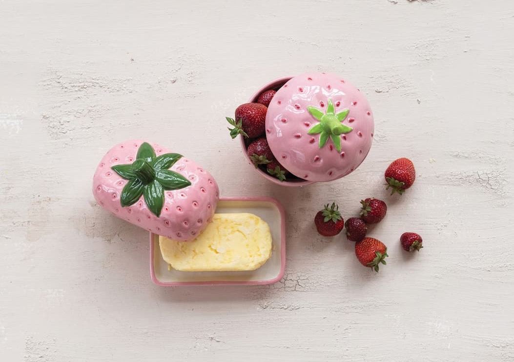 Overhead view of two pink strawberry-shaped ceramic containers on a light textured surface. One container is filled with fresh strawberries, while the other serves as a butter dish with a removable strawberry lid resting beside a block of butter. Scattered strawberries and glossy green leaf details create a playful, charming kitchen display.